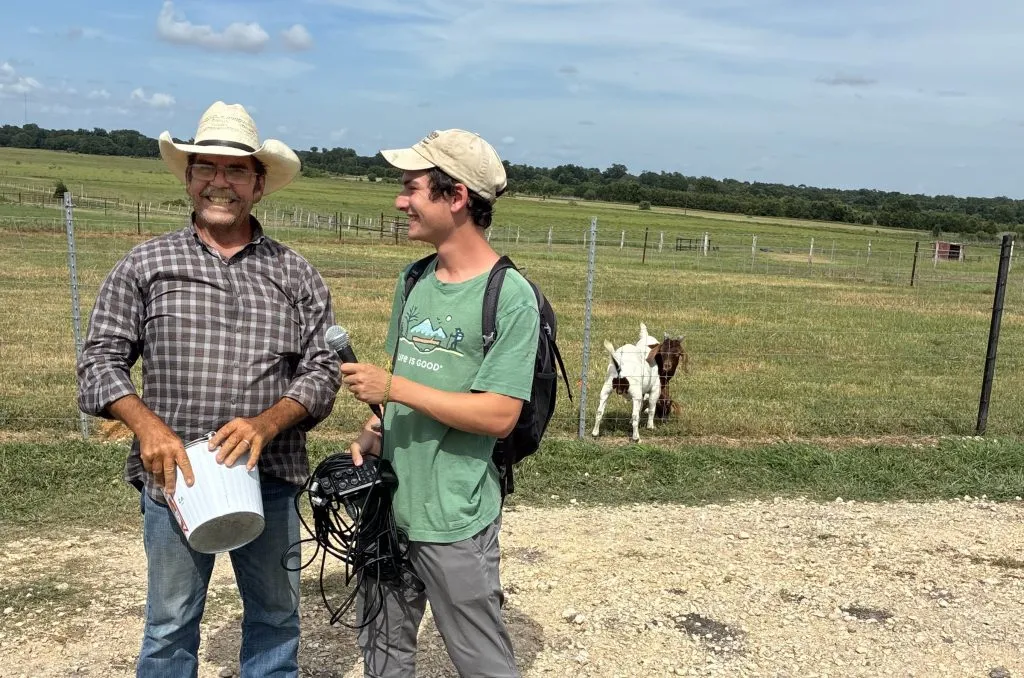Young man interviewing a rancher outside with goats in the background.